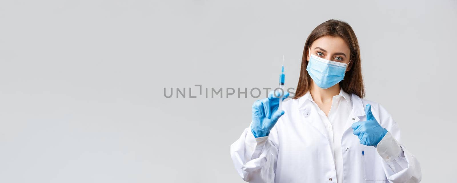 Covid-19, preventing virus, healthcare workers and quarantine concept. Determined young doctor in medical mask and rubber gloves, scrubs show thumbs-up and syringe with coronavirus vaccine.