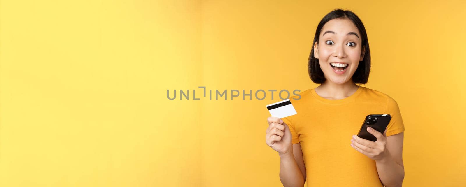 Online shopping. Cheerful asian girl holding credit card and smartphone, paying, order with mobile phone, standing over yellow background.