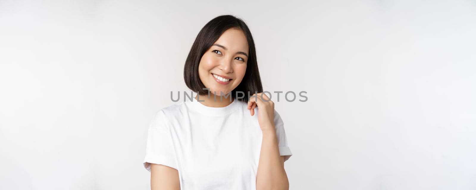 Portrait of cute coquettish woman laughing and smiling, looking aside thoughtful, thinking or imaging smth, standing in white t-shirt over studio background.