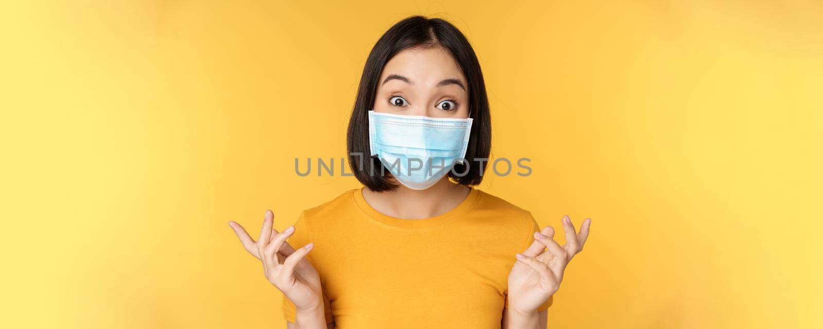 Covid-19 and medical concept. Close up portrait of asian woman in face mask, looking surprised and amazed at news, standing over yellow background.