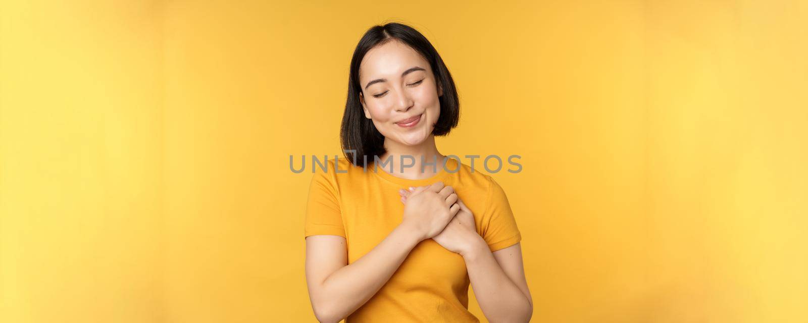 Romantic korean girl, asian woman holding hands on heart, smiling with care and tenderness, standing over yellow background.