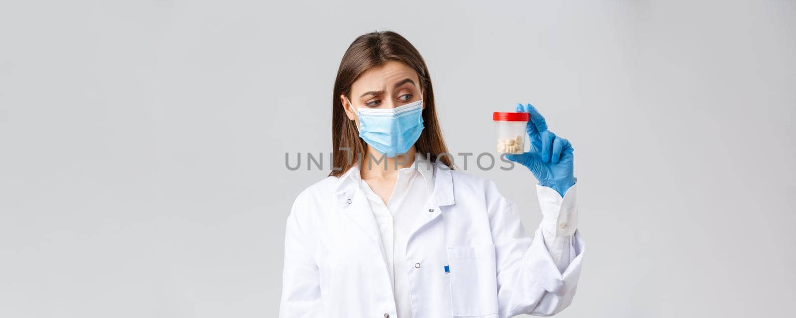 Covid-19, preventing virus, healthcare workers and quarantine concept. Unsure, curious doctor in scrubs and medical mask look intrigued and uncertain at container with medicine pills.