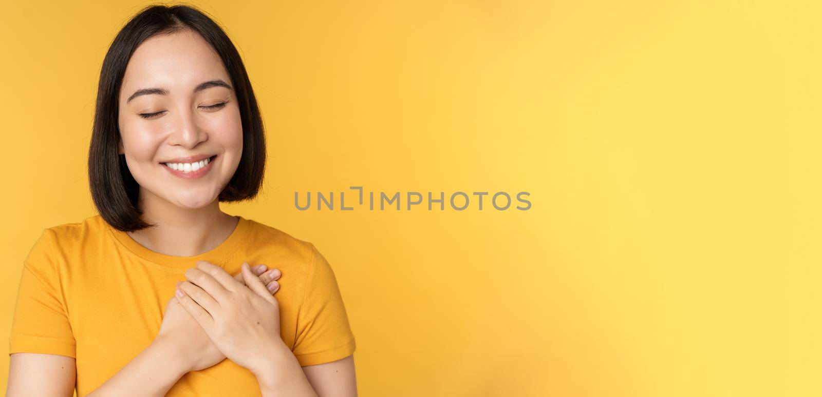 Beautiful asian woman, smiling with tenderness and care, holding hands on heart, standing in tshirt over yellow background by Benzoix