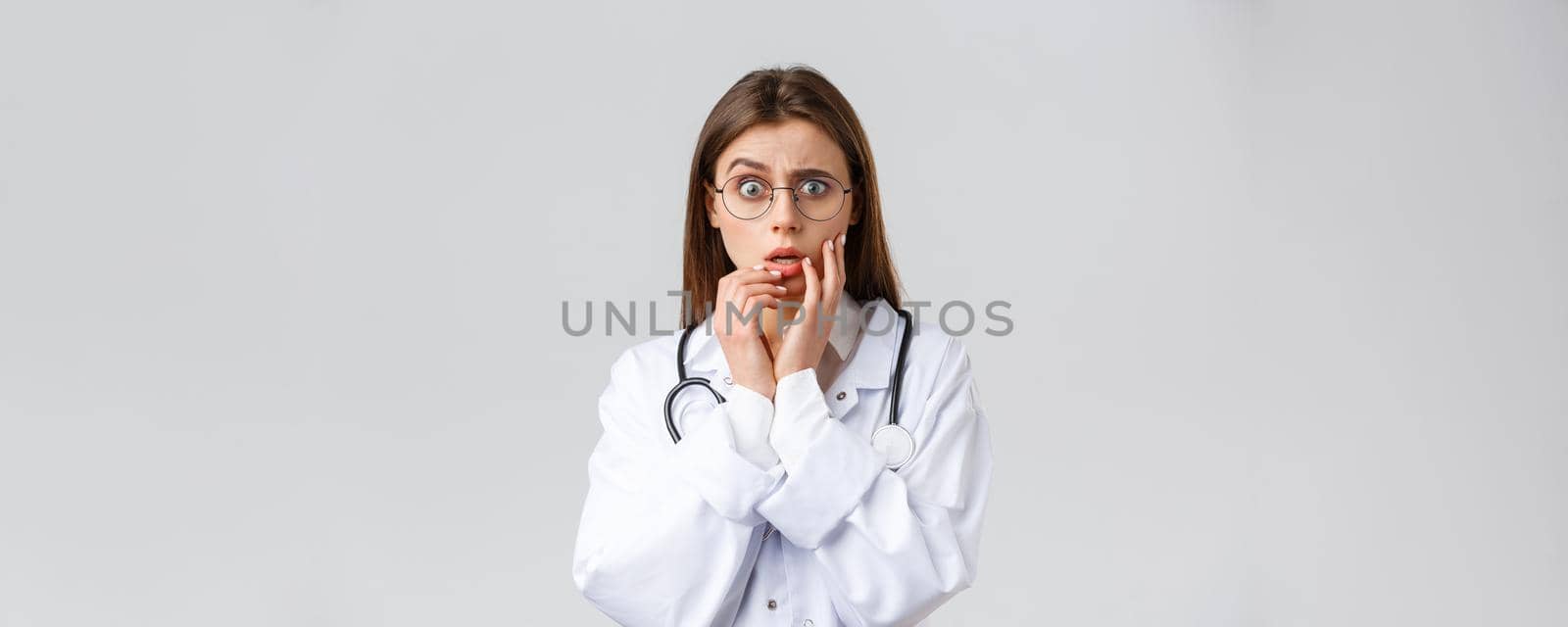 Healthcare workers, medicine, insurance and covid-19 pandemic concept. Shocked scared and concerned female doctor in white scrubs and glasses listening to scary news, gasping, hold hands near lips.