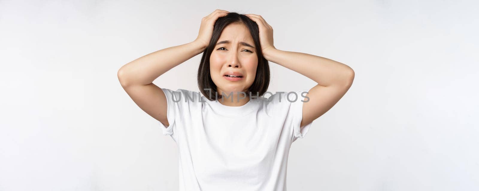 Desperate young korean woman holding hands on head, panicking, crying and standing distressed against white background by Benzoix