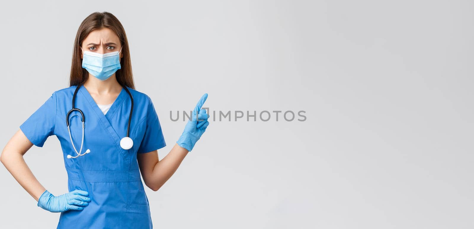 Covid-19, preventing virus, health, healthcare workers and quarantine concept. Skeptical or doubtful female nurse in blue scrubs and medical mask, gloves pointing right, frowning disbelief by Benzoix