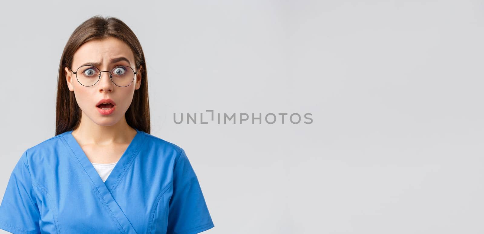 Healthcare workers, medicine and covid-19 pandemic concept. Confused and startled female nurse, doctor in blue scrubs and glasses, staring shocked, raise eyebrow questioned, grey background by Benzoix