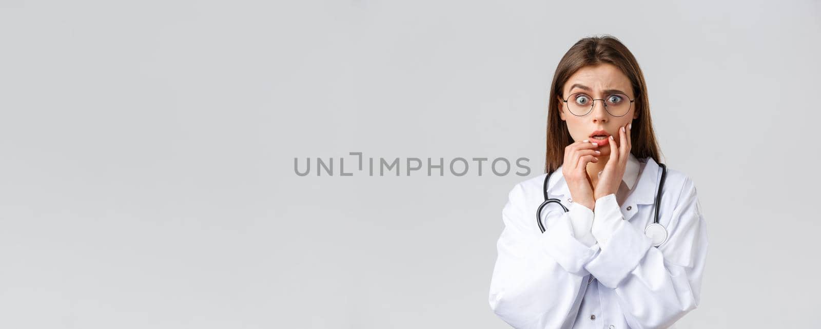 Healthcare workers, medicine, insurance and covid-19 pandemic concept. Shocked scared and concerned female doctor in white scrubs and glasses listening to scary news, gasping, hold hands near lips by Benzoix