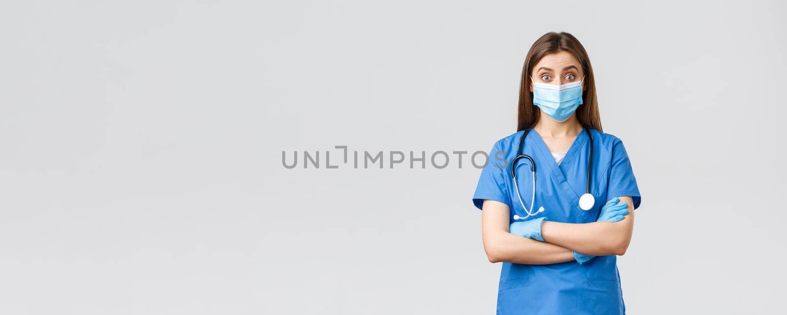 Covid-19, preventing virus, health, healthcare workers and quarantine concept. Amused female doctor or nurse in blue scrubs with stethoscope and personal protective equipment look intrigued.