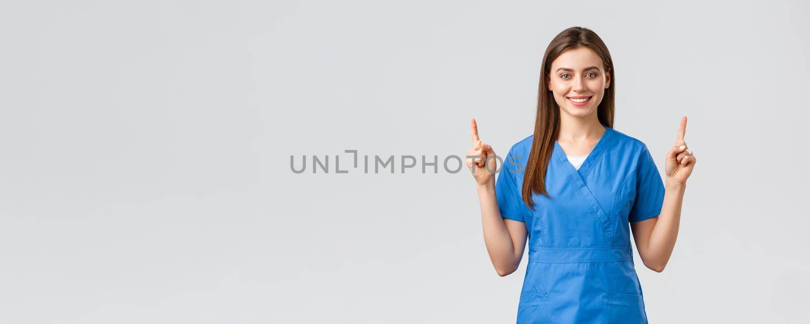 Healthcare workers, prevent virus, insurance and medicine concept. Attractive nurse or doctor in blue scrubs, pointing fingers up and smiling, advertise banner, medical pills, grey background.