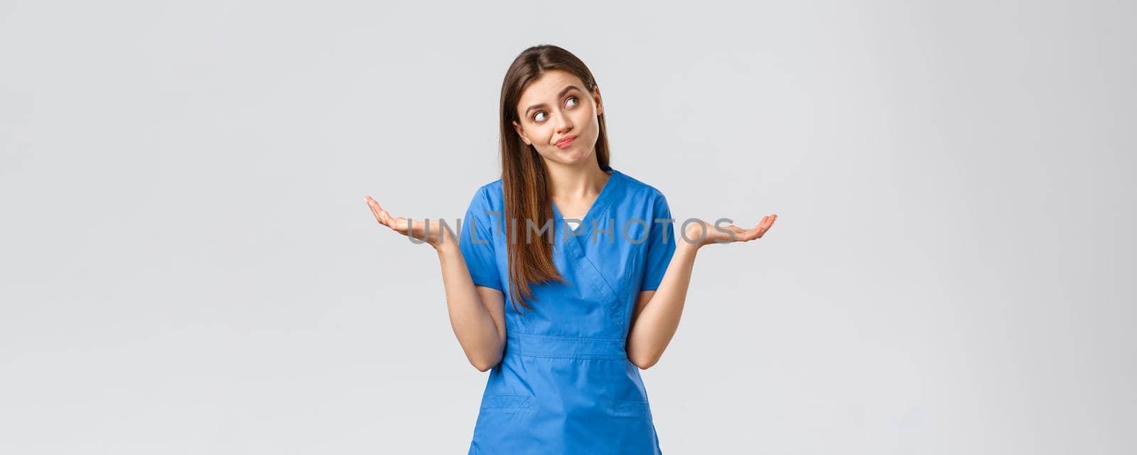 Healthcare workers, prevent virus, insurance and medicine concept. Indecisive and clueless attractive doctor, female nurse in blue scrubs, shrugging with hands sideways and smirk, look away.