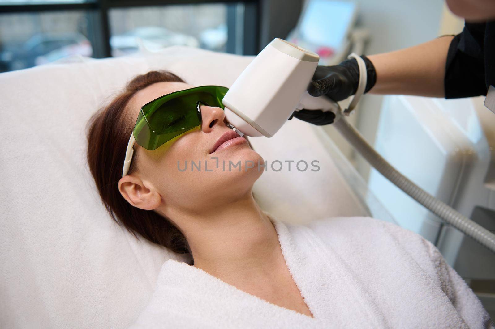 Doctor aesthetics beautician treats the face of a beautiful middle aged Caucasian woman with a professional modern laser epilator. Hardware cosmetology, beauty treatment, skin and body care concept