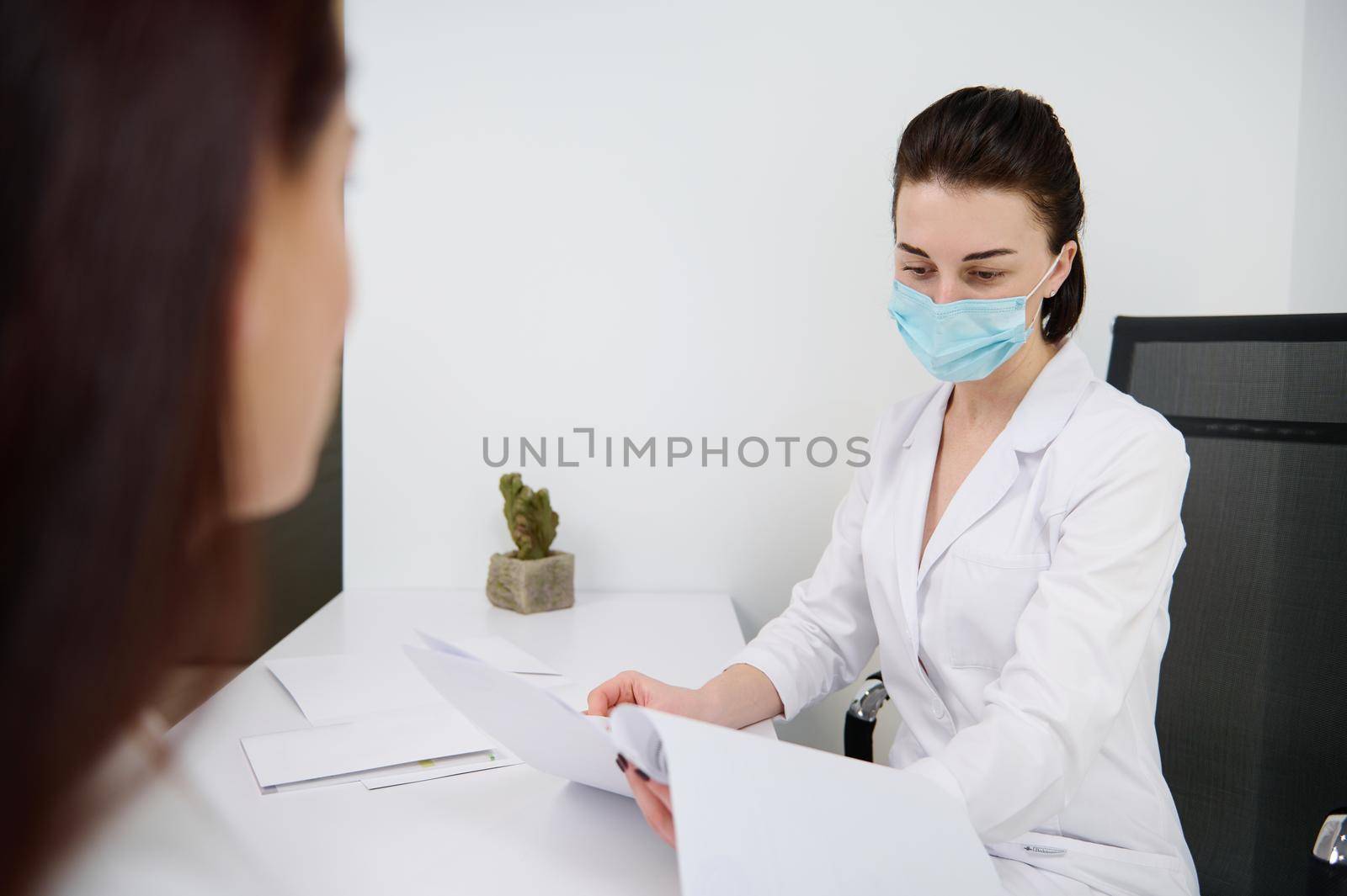 Woman at a doctor's appointment in a white office. Confident general practice cosmetologist beautician wearing medical protective mask reading medical documentation during examination her patient by artgf