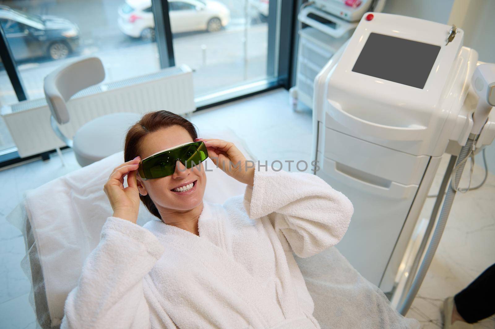 Charming woman smiles cheerfully while putting on UV protective goggles while lying on a medical massage couch, ready to get a beauty laser treatment in wellness spa clinic with modern equipments