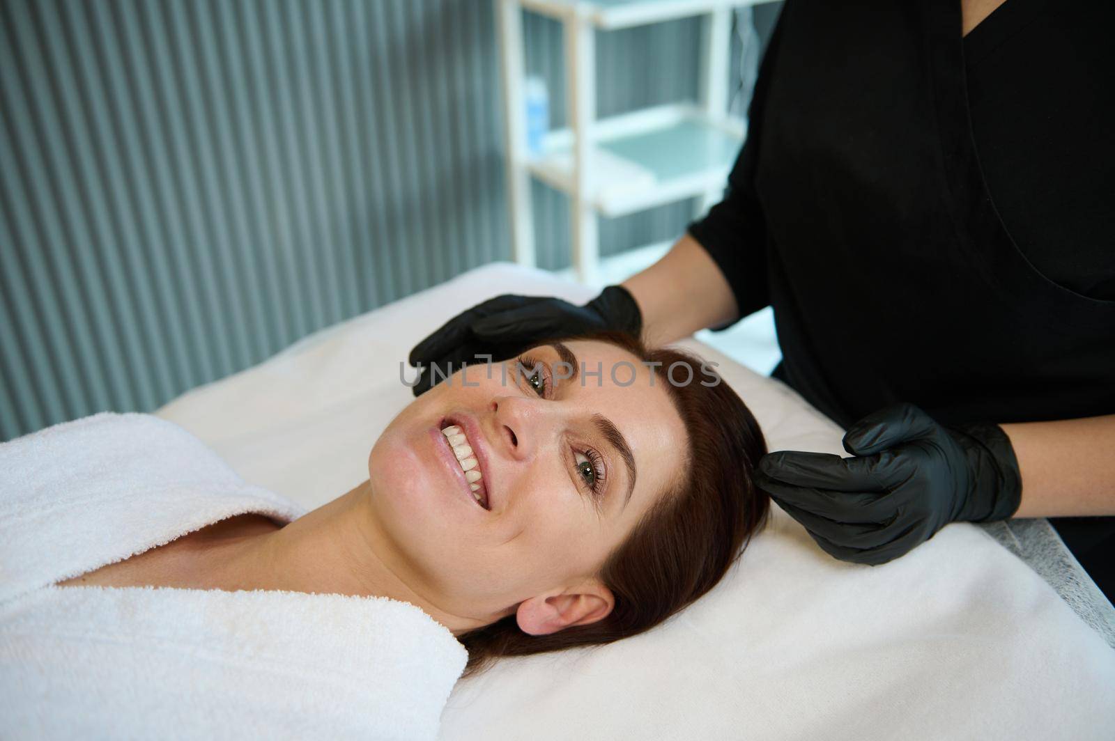 Happy and beautiful middle aged dark-haired European woman smiling with cheerful toothy smile while lying on a massage couch, enjoying beauty and wellness therapy in modern spa salon by artgf