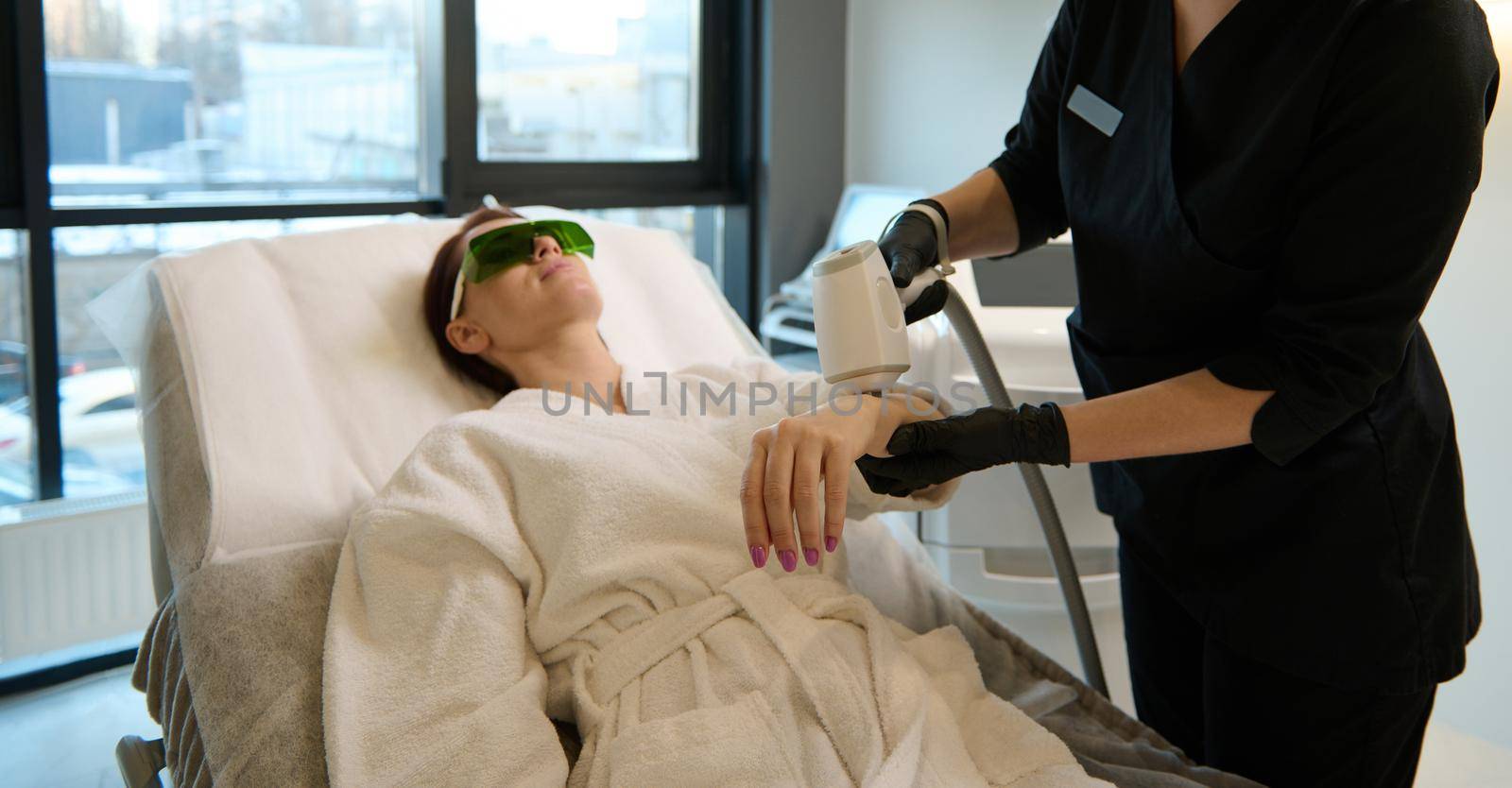 Cropped view of unrecognizable beautician performing a hair removal procedure on the hand and body of a Caucasian woman in bathrobe lying on a massage table and enjoying beauty treatment in spa salon