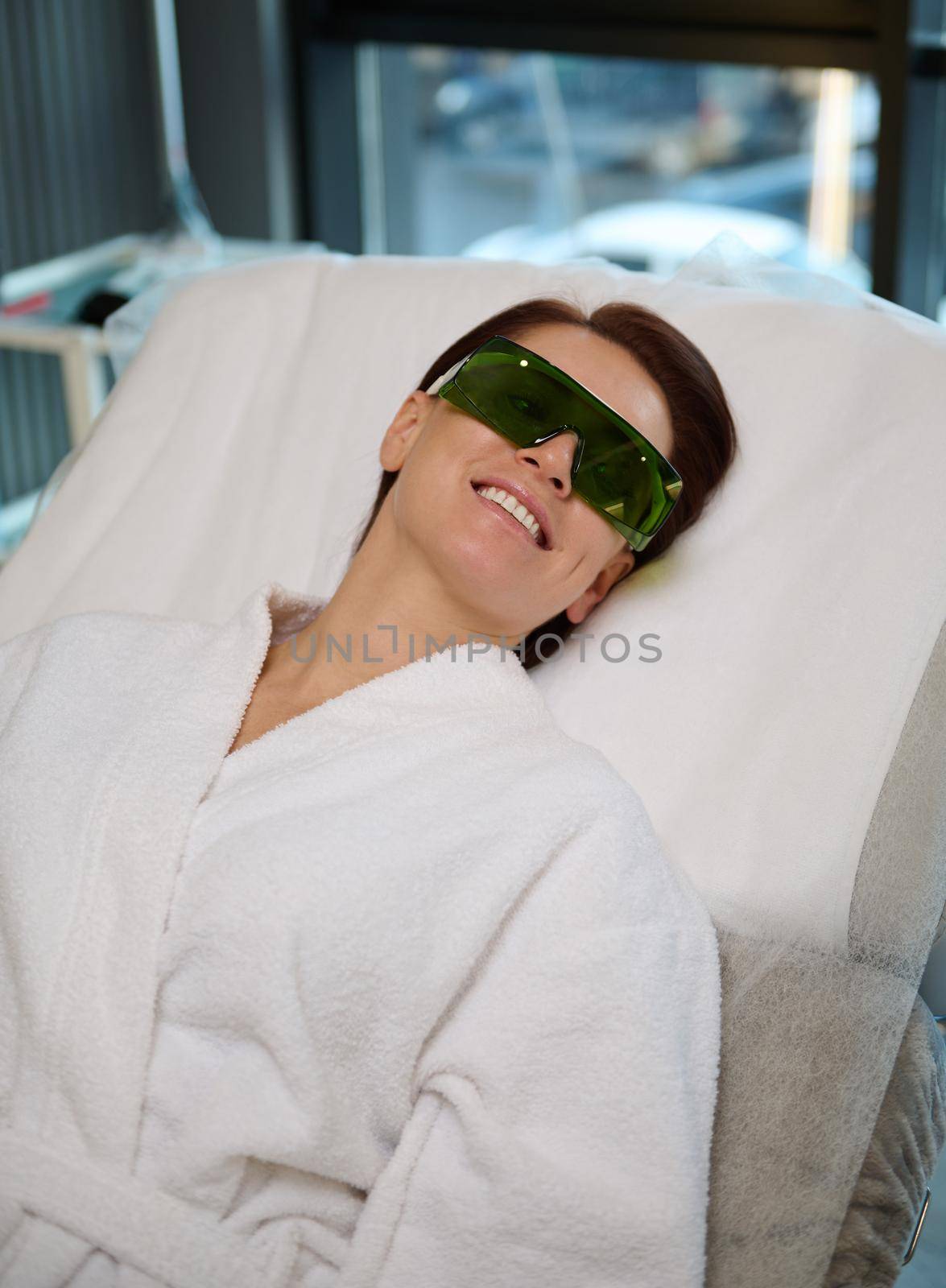 Charming middle aged woman, female patient with protective UV eye goggles, wearing a white terry bathrobe lying on a massage couch during wellness and body care procedures in modern spa resort