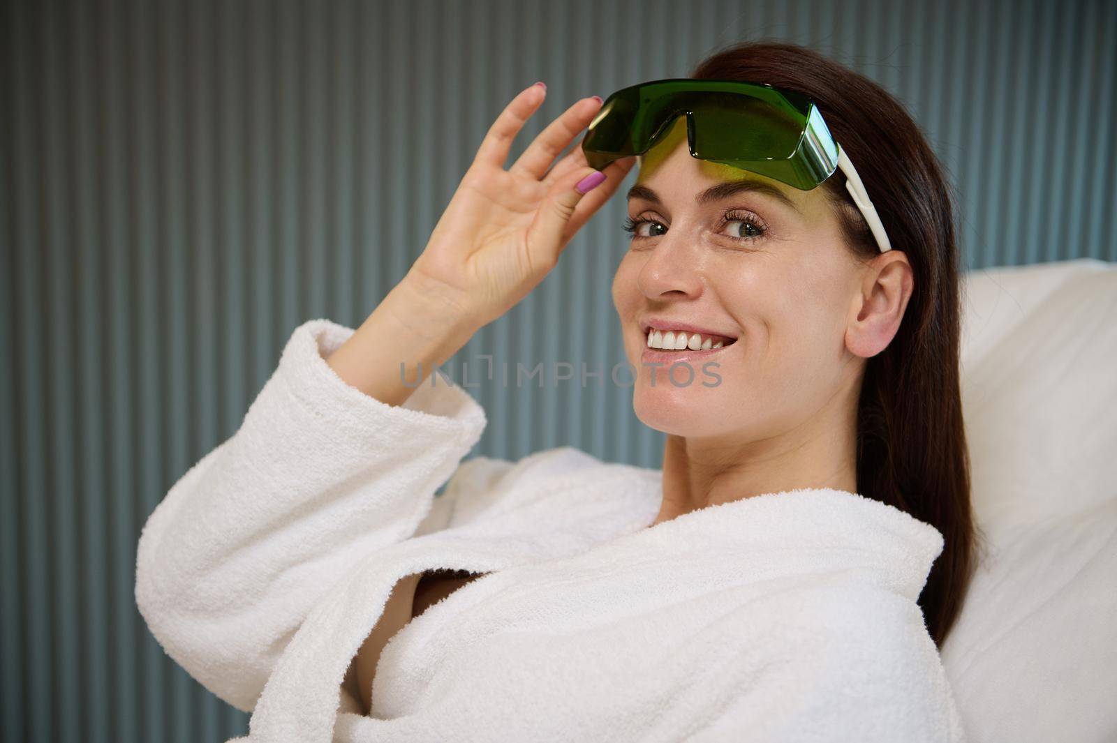 Attractive middle aged woman smiling with a beautiful toothy smile, looking at camera while raising protective uv goggles, lying on a massage couch in laser therapy room in modern luxury spa clinic