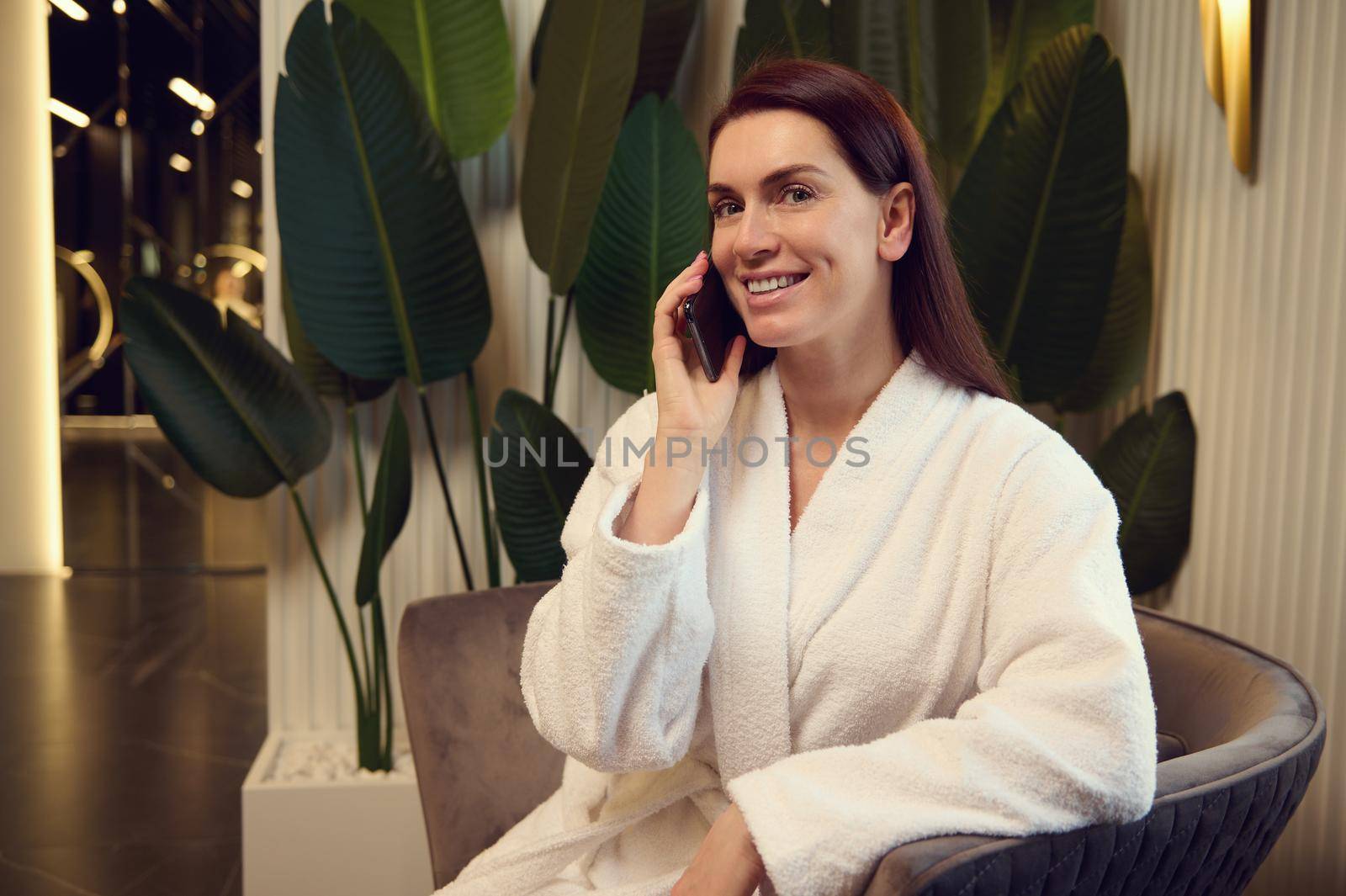 Attractive happy Caucasian woman in white terry bathrobe talking on mobile phone sitting in the spa lounge area after receiving wellness and beauty treatment in modern luxury spa clinic