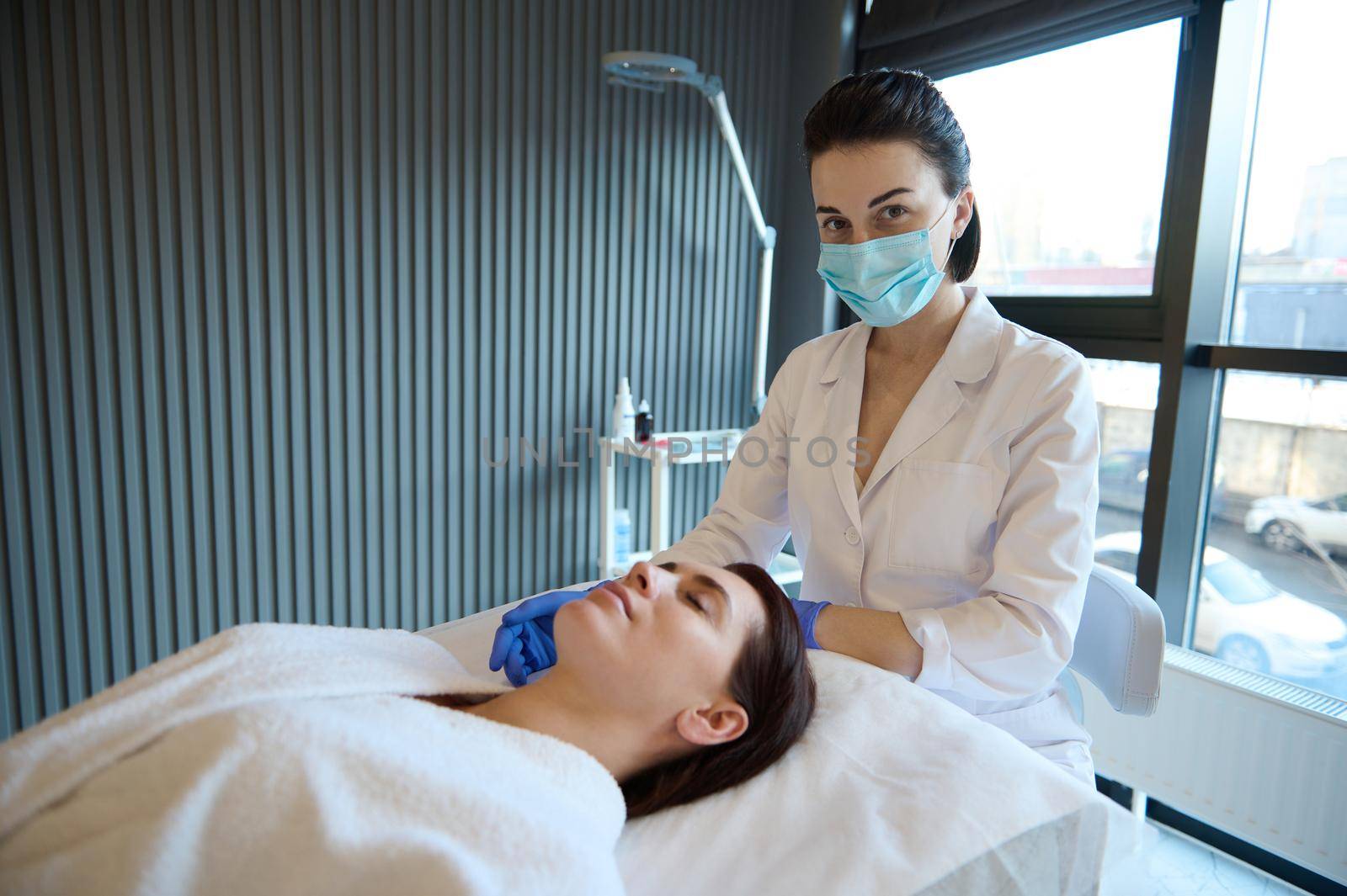 Young cosmetologist performs wellness procedures on the face of a relaxed middle-aged woman lying on a massage couch in a cosmetology spa clinic. Anti-age treatment and professional skin care concept