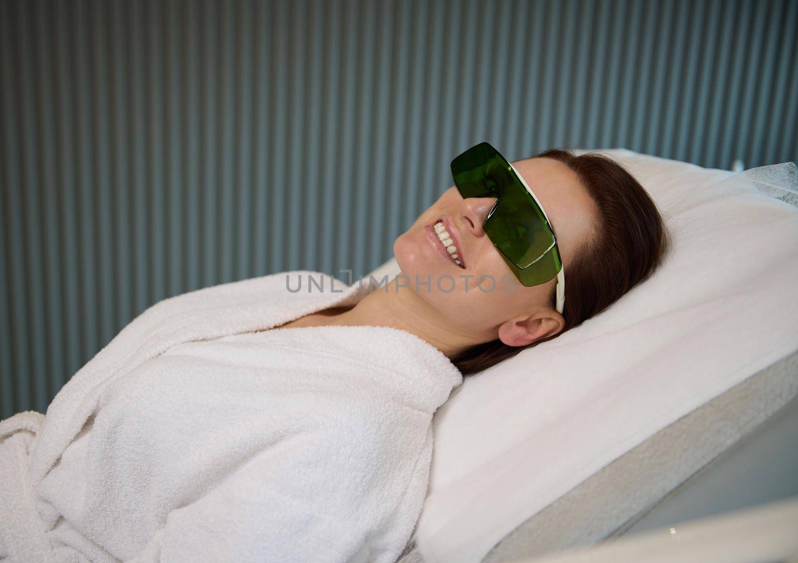 Charming pleased middle aged woman in protective UV goggles lying on a medical massage couch in spa salon and smiles with beautiful toothy smile, enjoying wellness procedures. Cosmetology concept by artgf