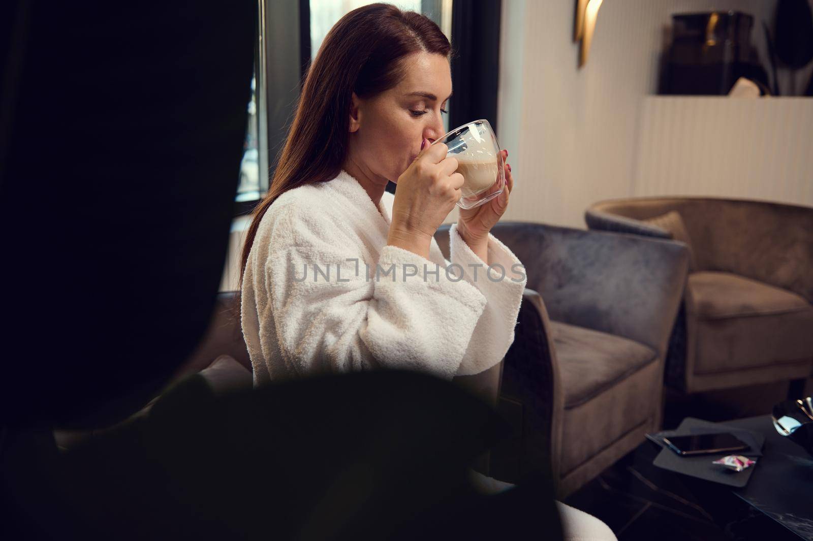 Beautiful woman drinking cappuccino, sitting on an armchair in a spa salon lounge area. Attractive relaxed European pretty woman enjoying a coffee break while visiting a luxury wellness spa clinic. by artgf