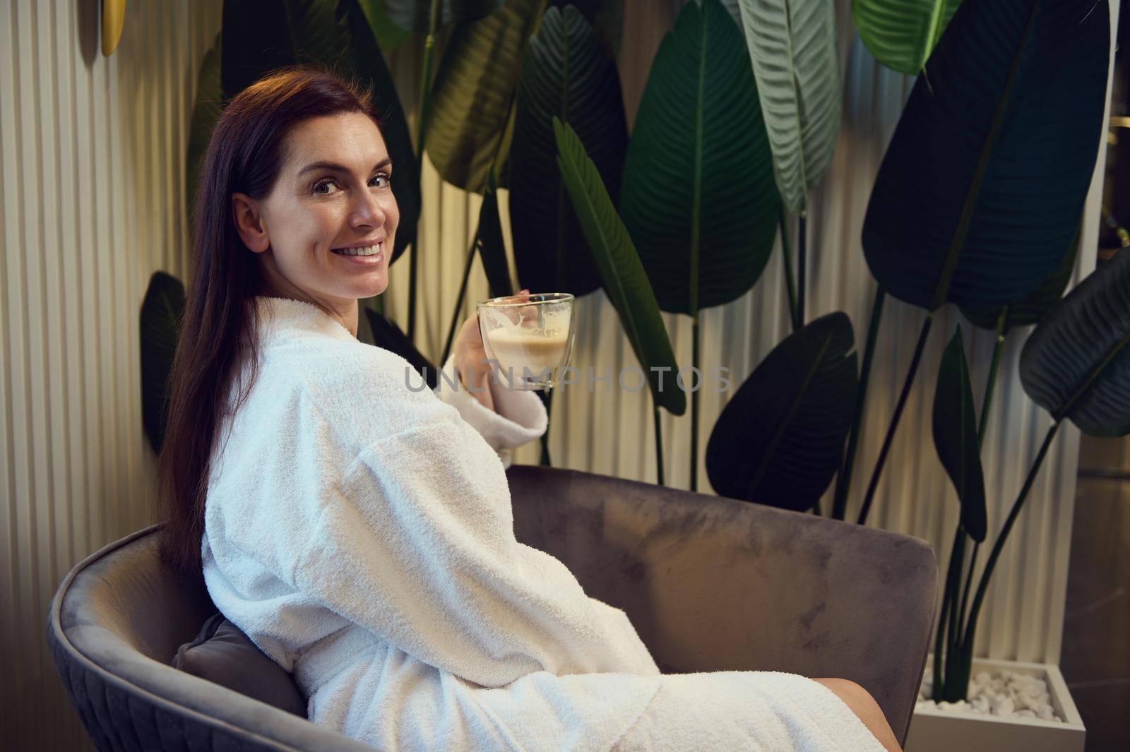 Charming Caucasian middle aged woman in white bathrobe enjoying coffee break, sitting on an armchair in the lounge of a modern spa clinic, smiles cheerfully looking at camera