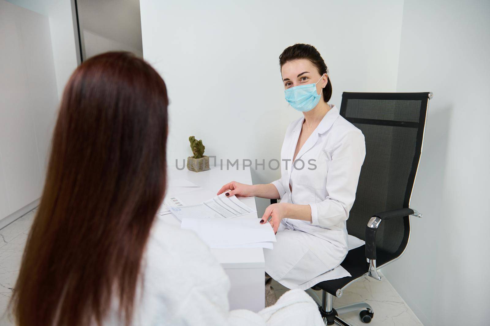 Woman at a doctor's appointment in a white office. Confident general practice cosmetologist beautician wearing medical protective mask reading medical documentation during examination her patient