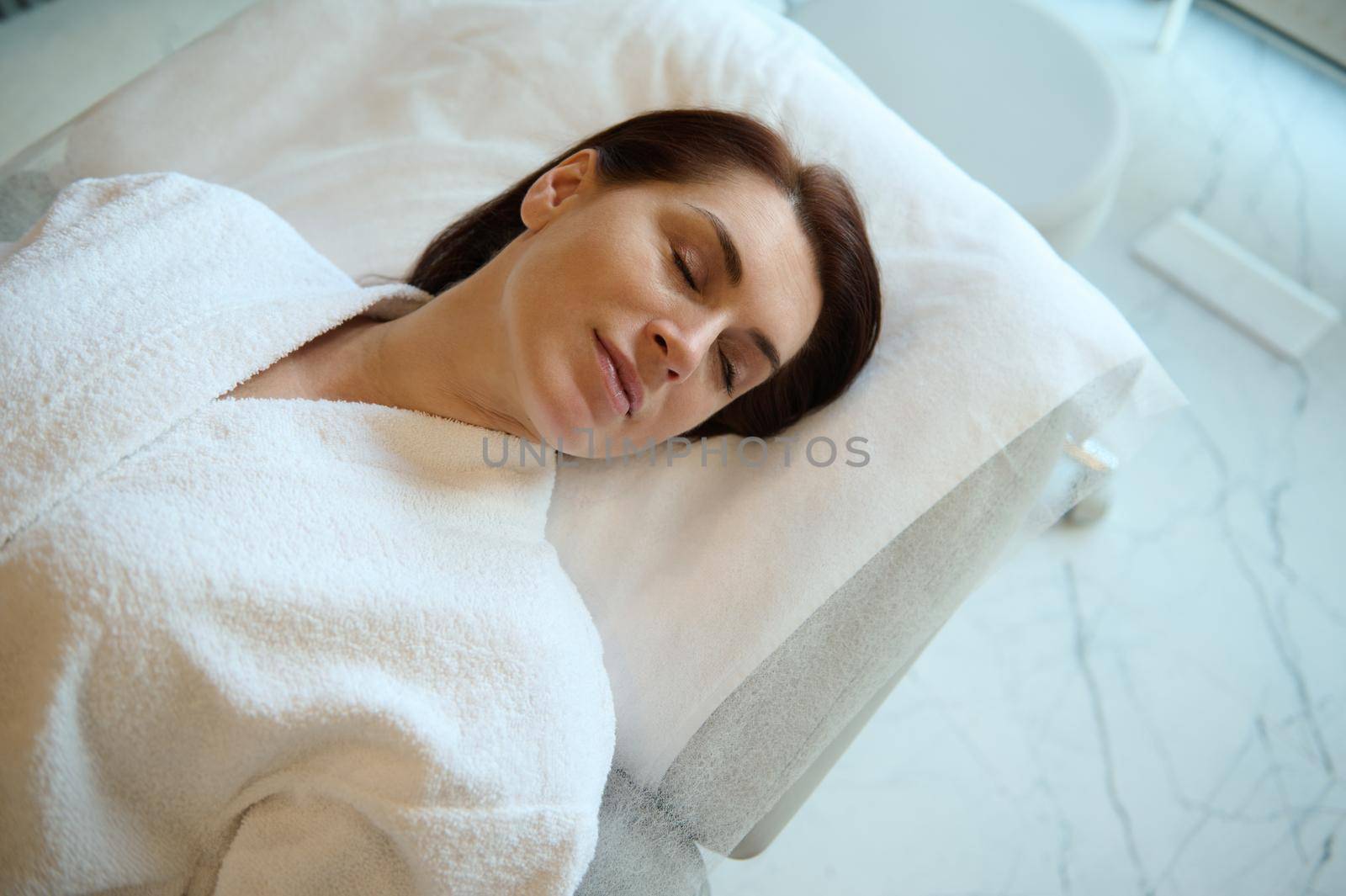 Overhead view of a beautiful Caucasian woman with perfect glowing skin relaxing on a massage table in a spa room, while receiving a professional skin and body care treatment in wellness spa clinic