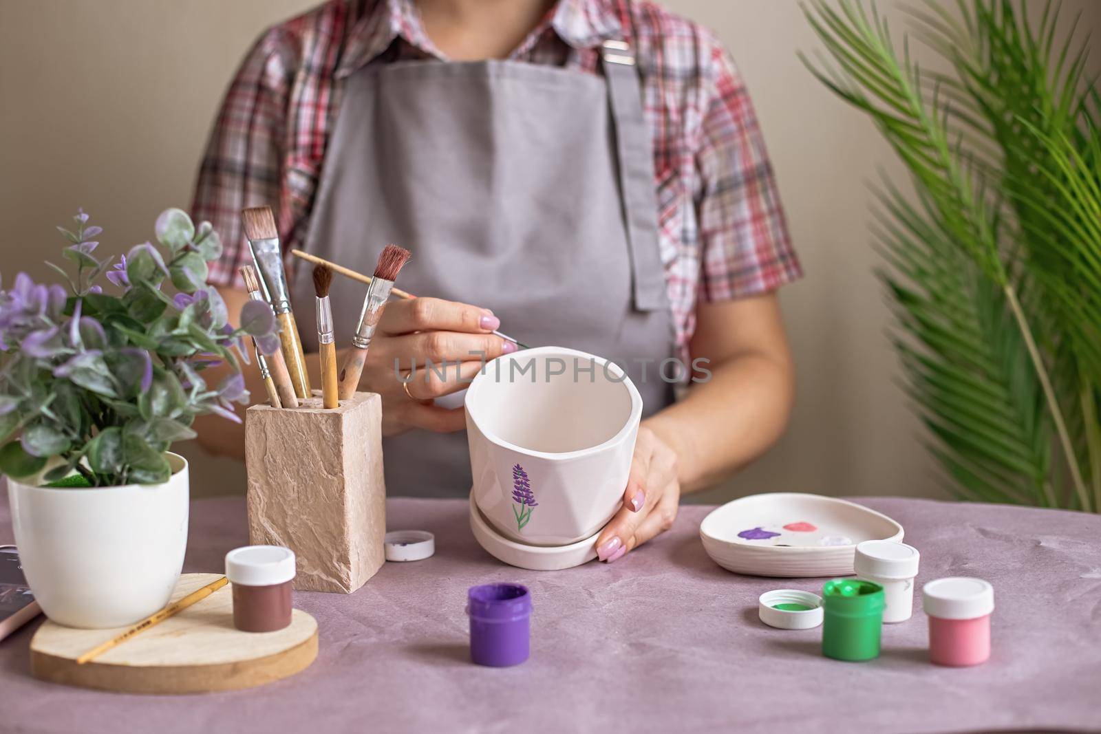 A woman in a gray apron and a plaid shirt, sits at a table, paints a white ceramic flower pot, in daylight. On the table is purple kraft paper, brushes, jars of paints. No face