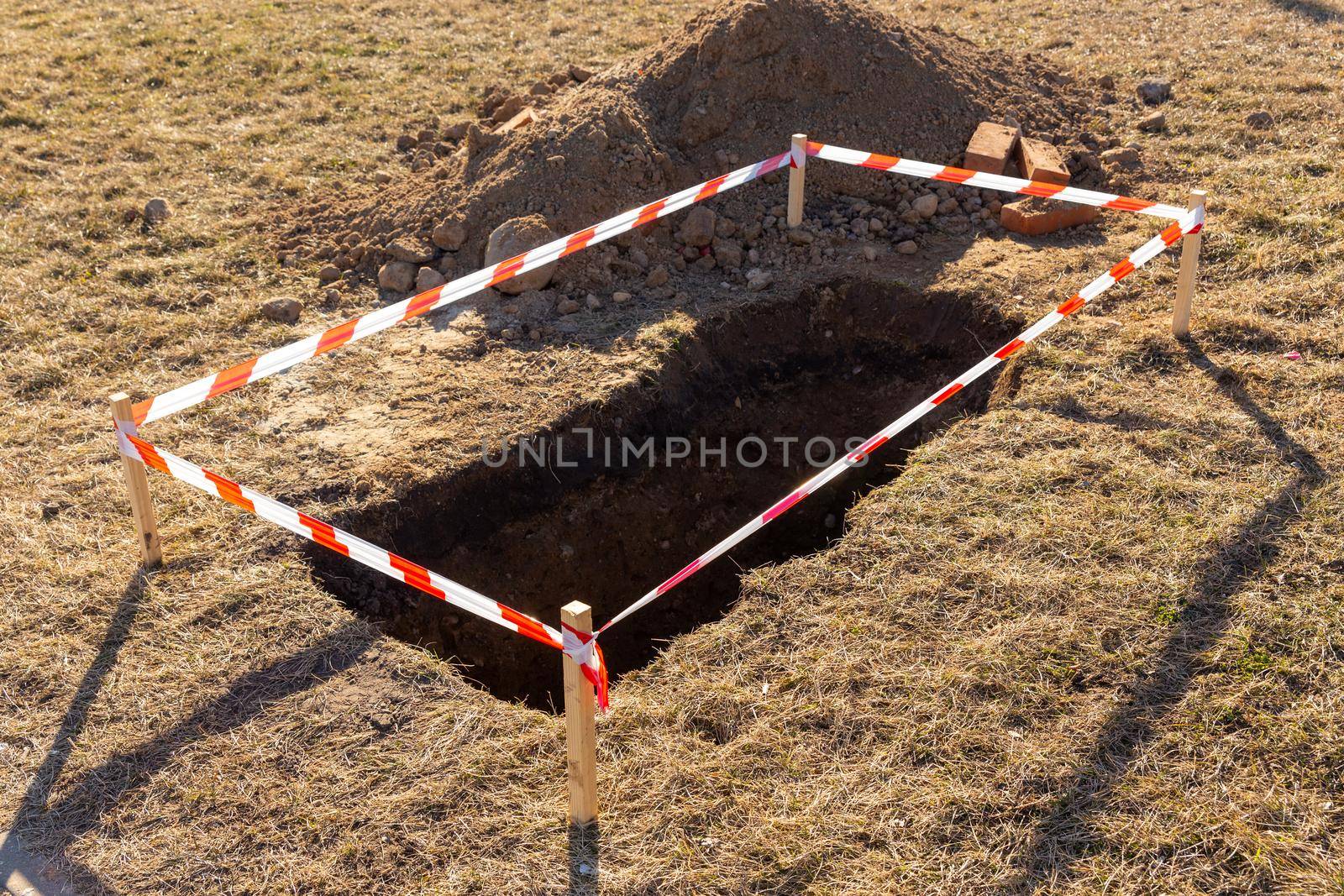 Archaeological excavations in the middle of the field. A small hole in the ground is fenced with red and striped construction tape. Ditch in the ground. Construction works.