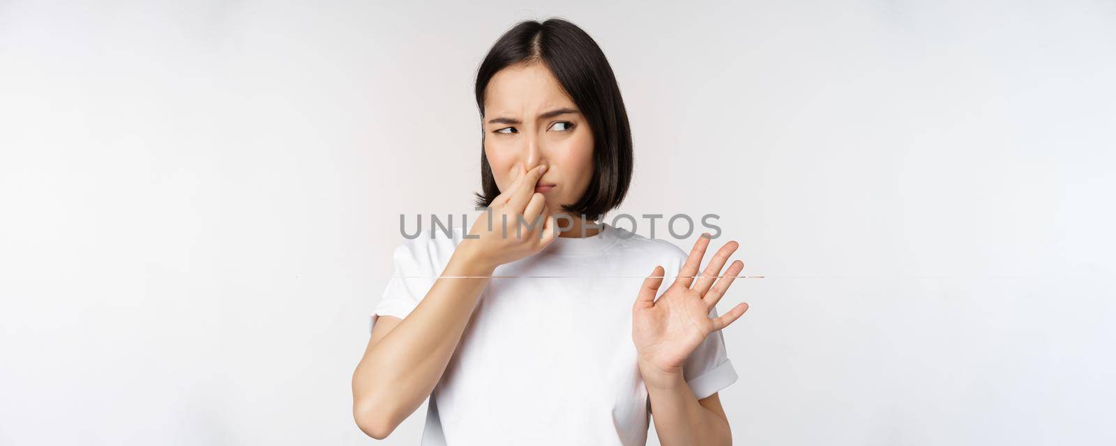 Asian girl looks disgusted, rejecting product with bad smell, shut nose from aversion and cringe, standing against white background.