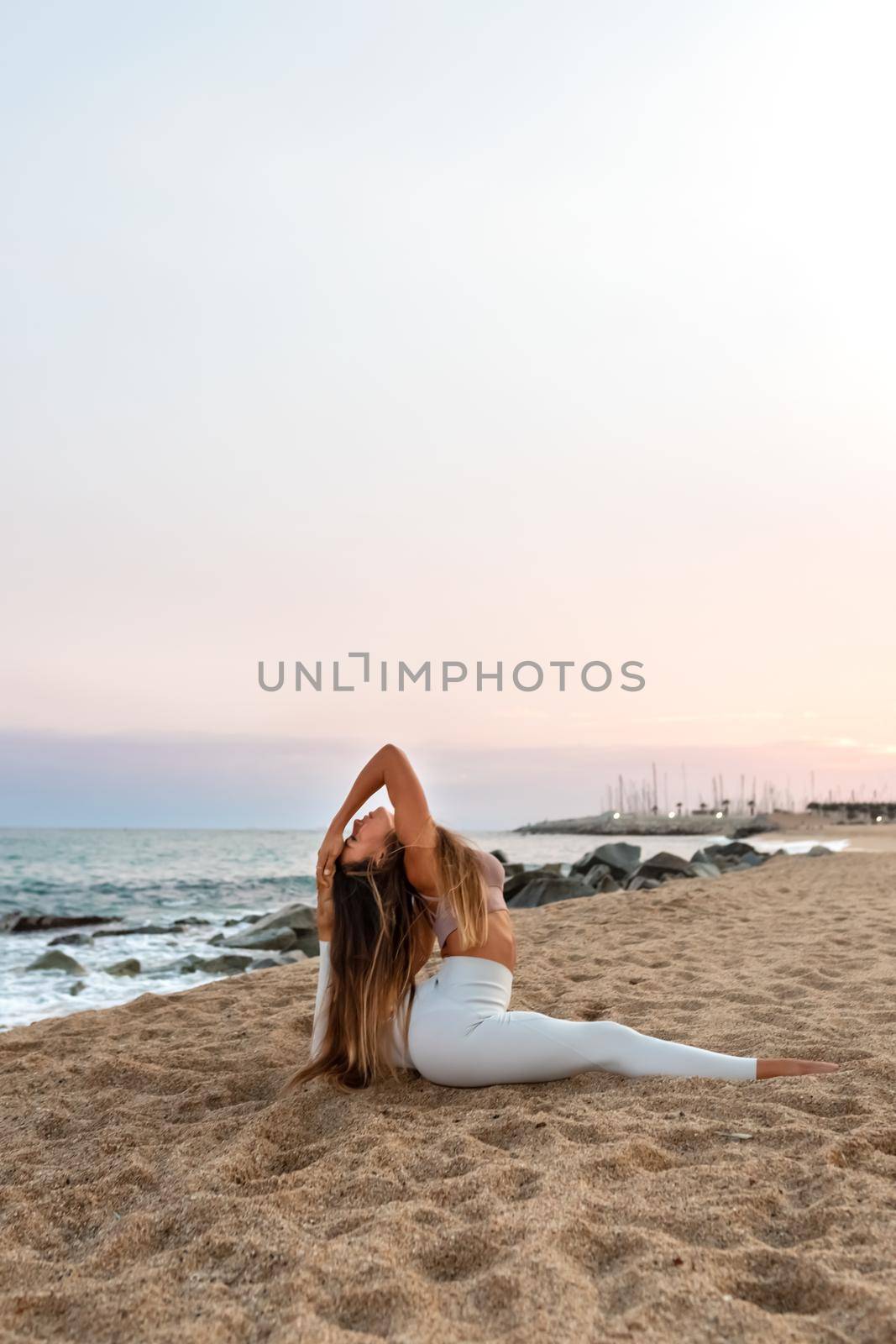 Young flexible caucasian woman practicing yoga doing splits on the beach at sunset. Copy space. Vertical image. Spirituality and healthy lifestyle concept.