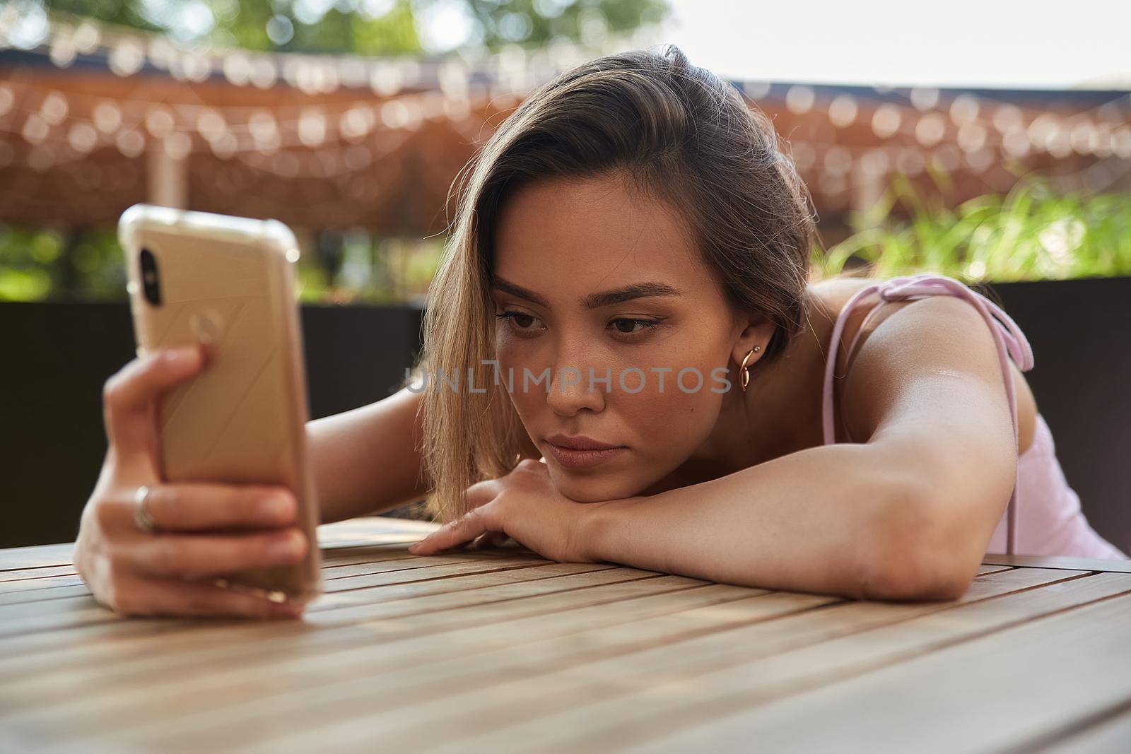 young asian pretty woman with long hair lays her head on her arms and table, holding phone in hand. beautiful pensive melancholic lady laying at cafe, using smartphone. lifestyle portrait