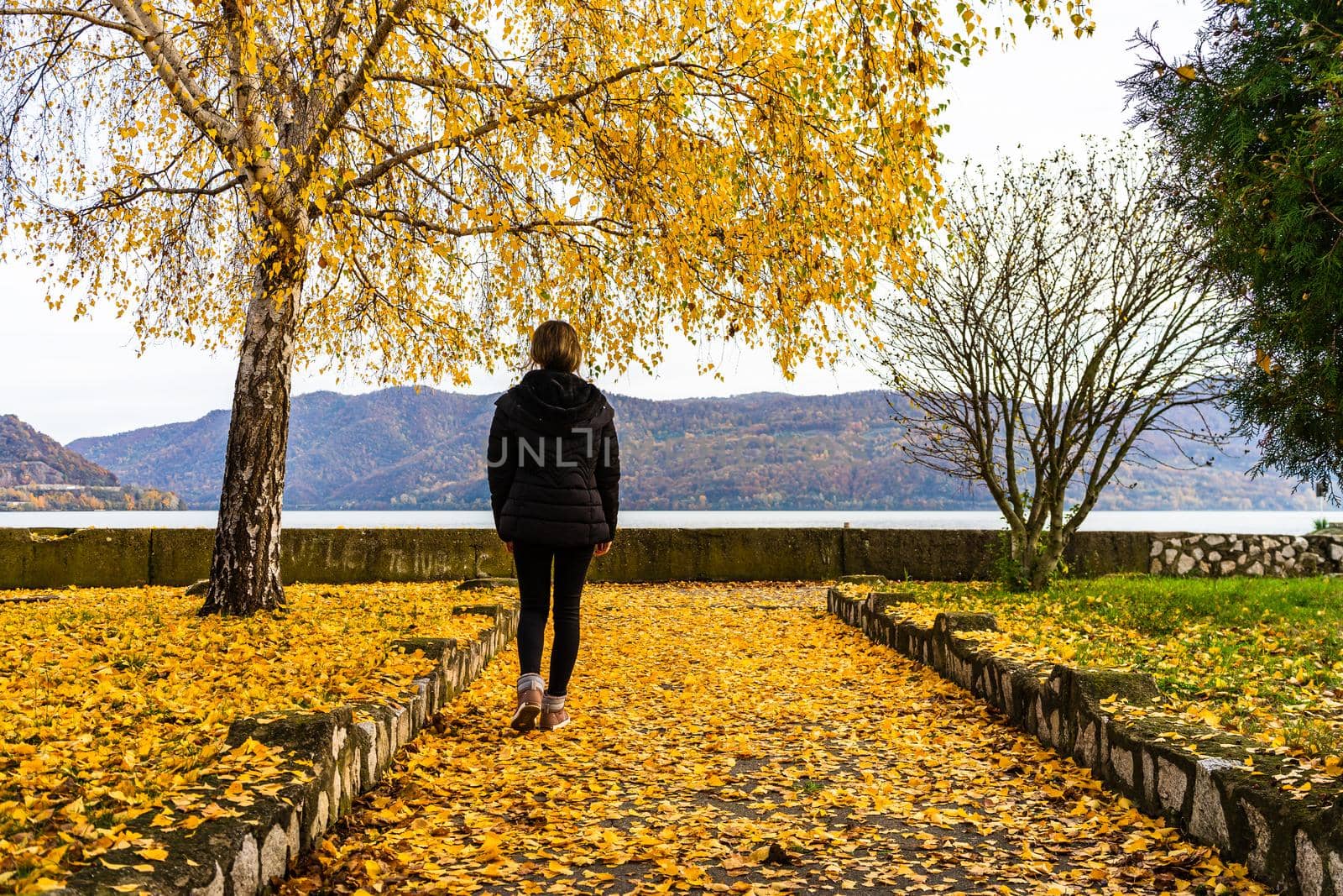Autumn leaves fallen on alone woman walking on the autumn alley. Autumn landscape, orange foliage in a park in Orsova, Romania, 2020
