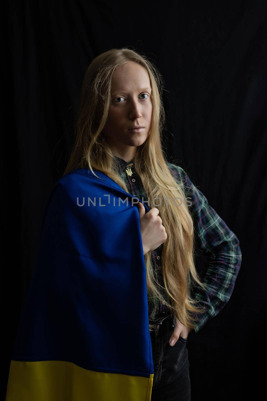 Woman Ukrainian blonde during the war with the state yellow blue flag and coat of arms on neck on isolated black background at studio. Russia attacked Ukraine on February 24, 2022. Concept - Stop war