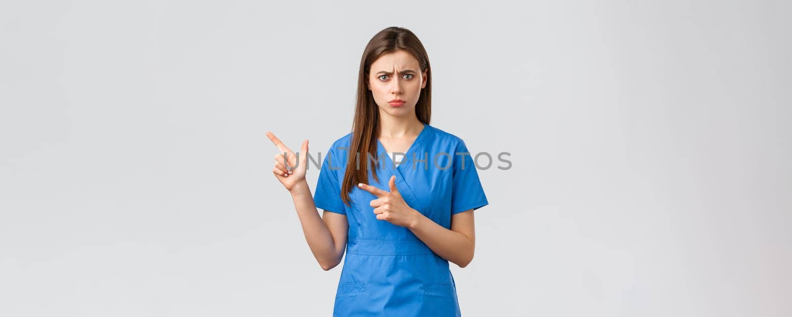 Healthcare workers, prevent virus, insurance and medicine concept. Concerned and alarmed young female doctor or nurse in blue scrubs frowning anxious, pointing fingers left at important info.