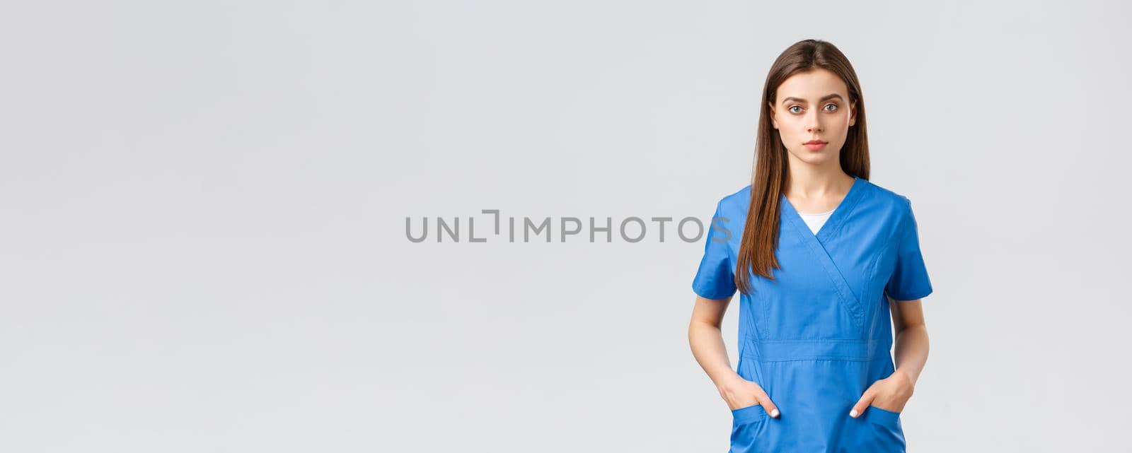 Healthcare workers, prevent virus, insurance and medicine concept. Serious-looking, confident young medical worker, nurse or doctor in blue scrubs hold hands in pockets and looking camera by Benzoix