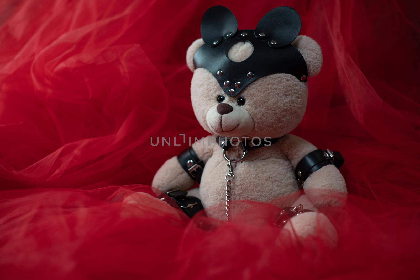 a toy teddy bear dressed in leather belts and a mask, an accessory for BDSM games against a beautiful red fabric by Rotozey