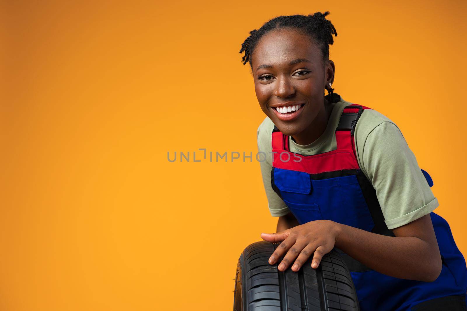 Portrait of smiling black female mechanic posing with new car tyres in yellow studio