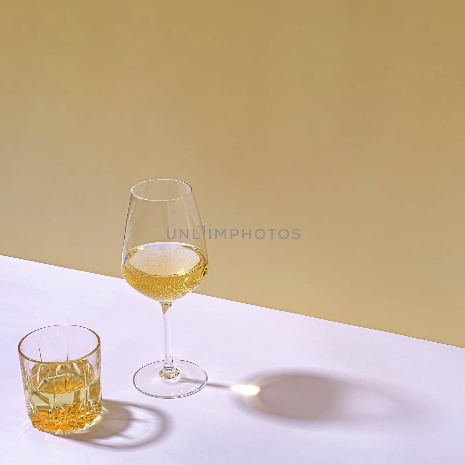 Tumbler of whiskey and glass of white wine side lit with shadow on a white table indoors with copy space