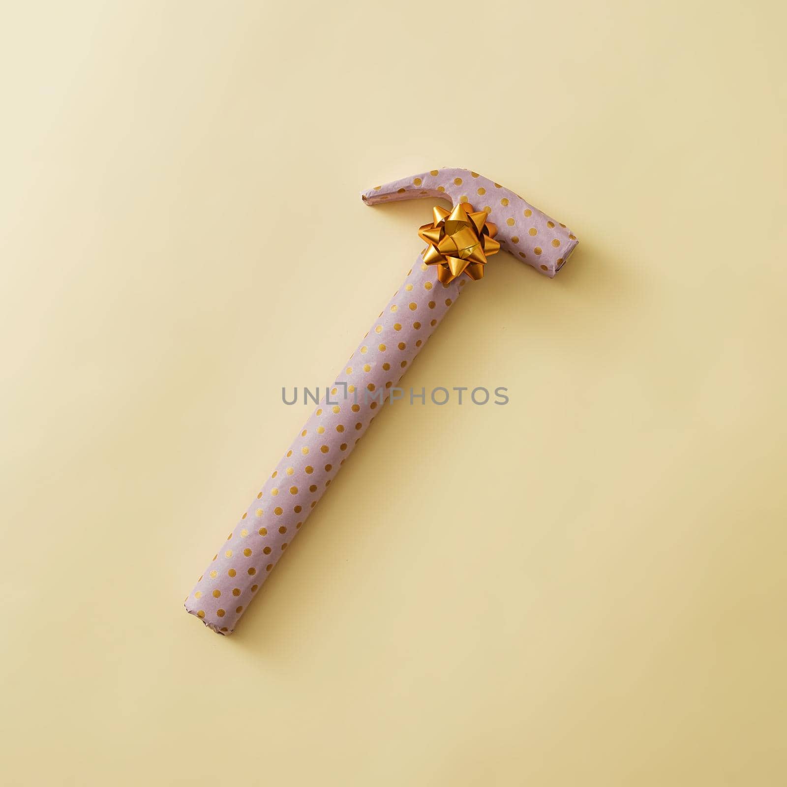 Gift wrapped hammer in yellow polka dot paper decorated with a gold ribbon bow over a yellow background with copy space