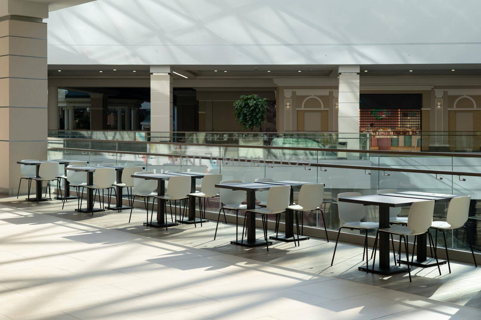 A row of tables with white chairs for visitors to the food court of a modern shopping center. People admire the beautiful view during the meal.