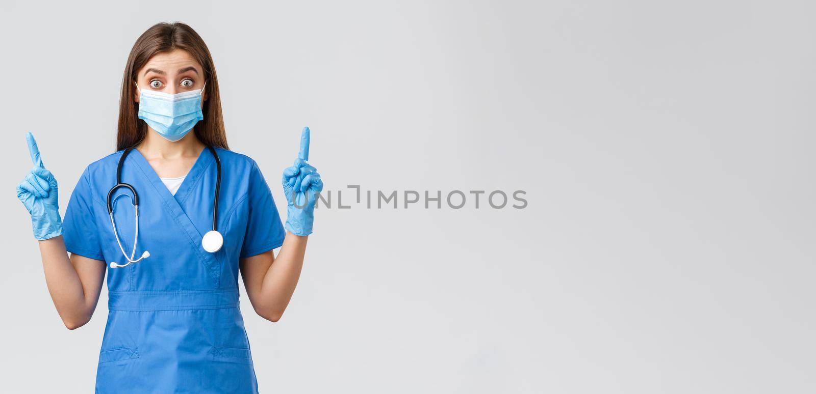 Covid-19, preventing virus, health, healthcare workers concept. Surprised or curious female nurse in blue scrubs, personal protective equipment, pointing fingers up, found out interesting news.