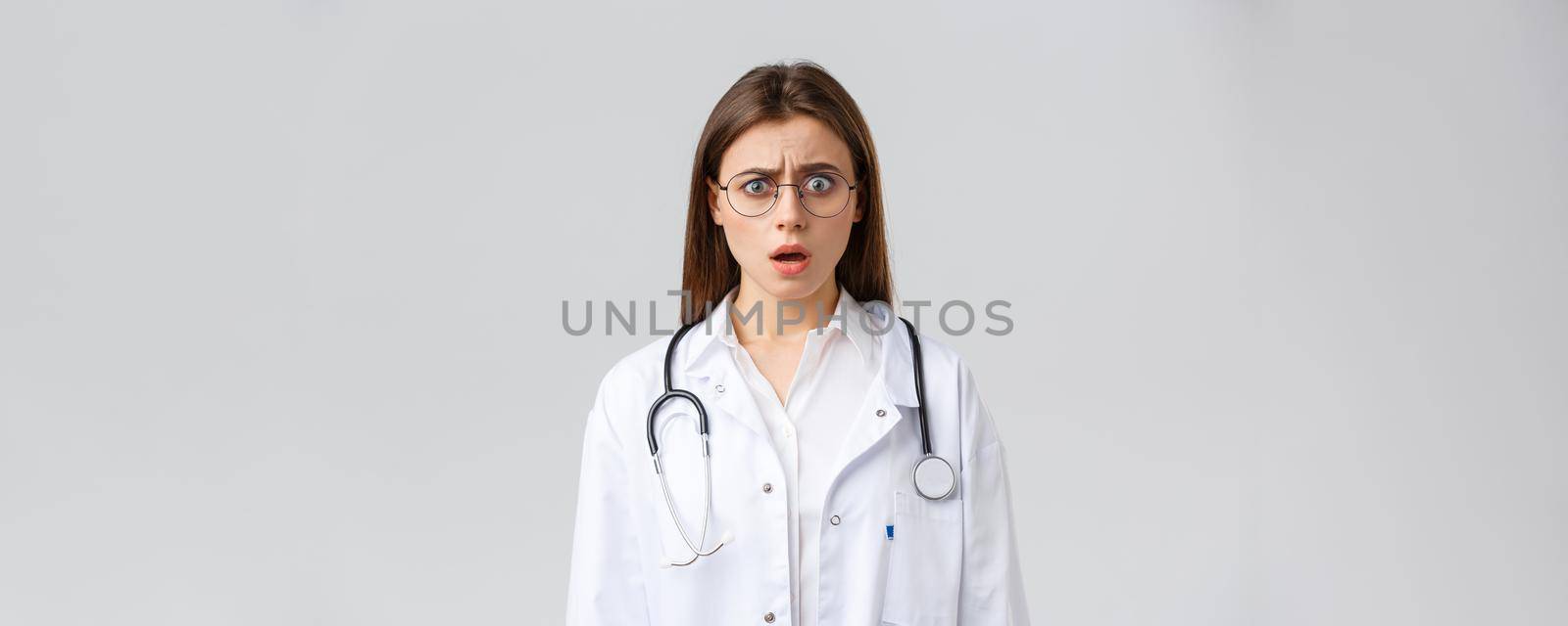 Healthcare workers, medicine, insurance and covid-19 pandemic concept. Shocked nervous female doctor in white scrubs and glasses, doctor gasping and staring concerned camera.