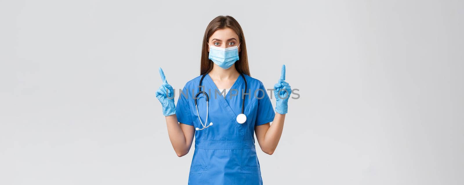 Covid-19, preventing virus, health, healthcare workers concept. Serious and confident female nurse in blue scrubs, medical mask PPE, pointing fingers up, inform patients how prevent corona infection.