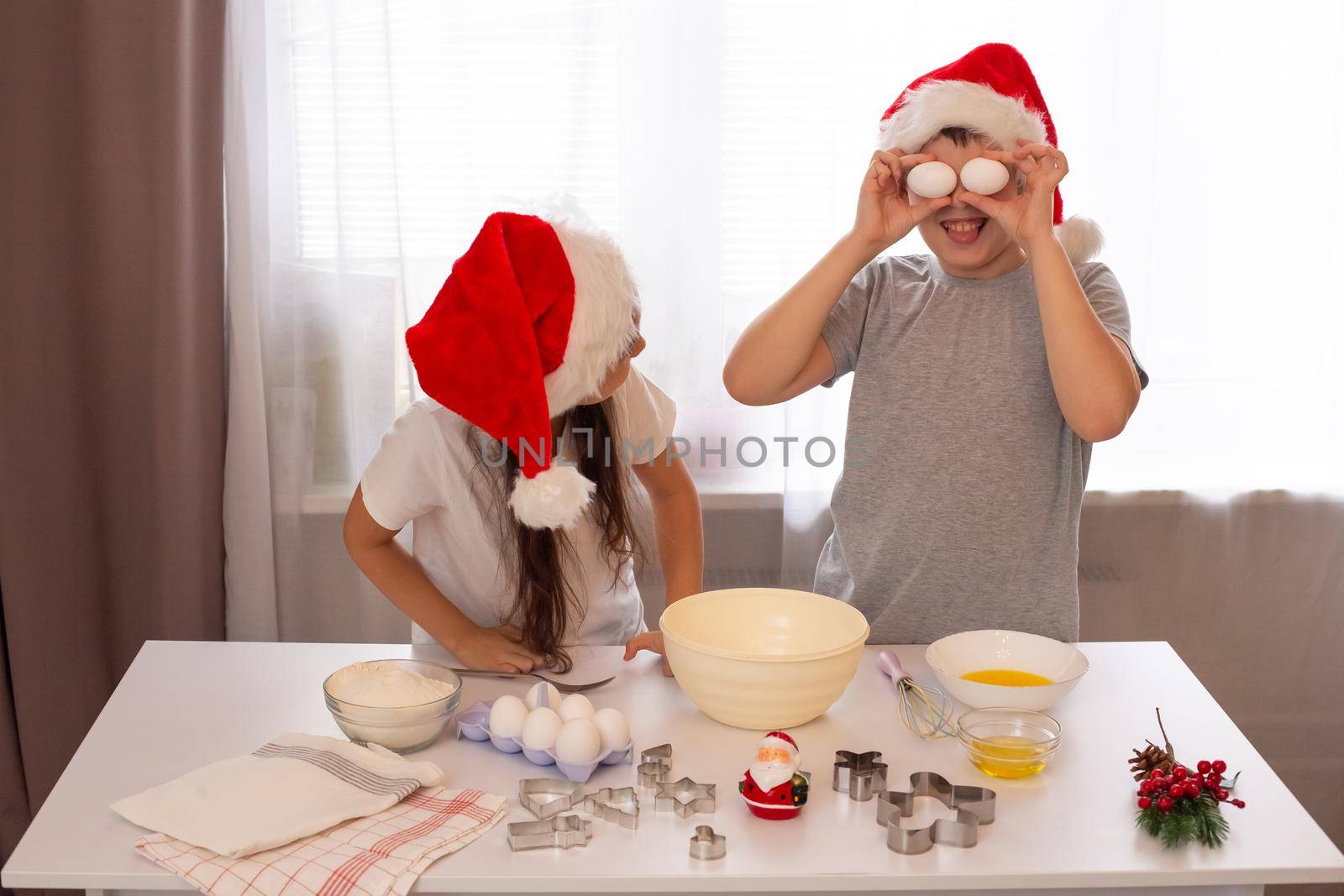 Brother and sister in red caps and light-colored clothes are having fun and preparing Christmas cookies at a white table. Boy holds two white eggs and laughs
