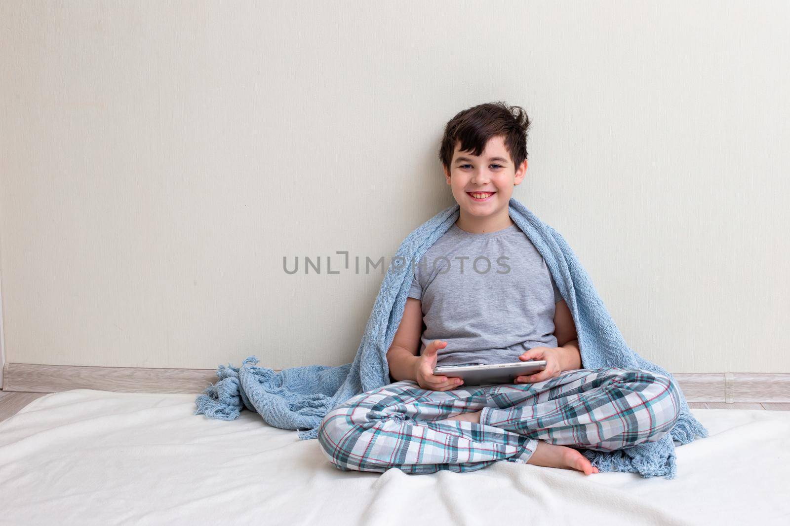 A handsome smiling boy, a teenager in gray pajamas, in a blue knitted plaid sits against a light wall, on the floor, holds a digital tablet, looks at the camera. Copy space