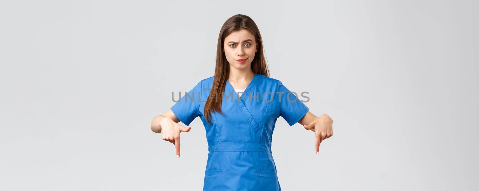 Healthcare workers, prevent virus, covid-19 test screening, medicine concept. Skeptical, disappointed female nurse or doctor in blue scrubs pointing fingers down, smirk unsatisfied, being doubtful.