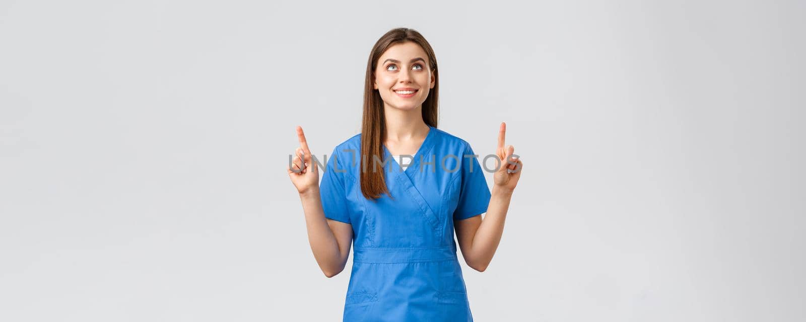 Healthcare workers, prevent virus, covid-19 test screening, medicine concept. Attractive female nurse or doctor in blue scrubs, pointing fingers and looking up with pleased smile, reading banner.